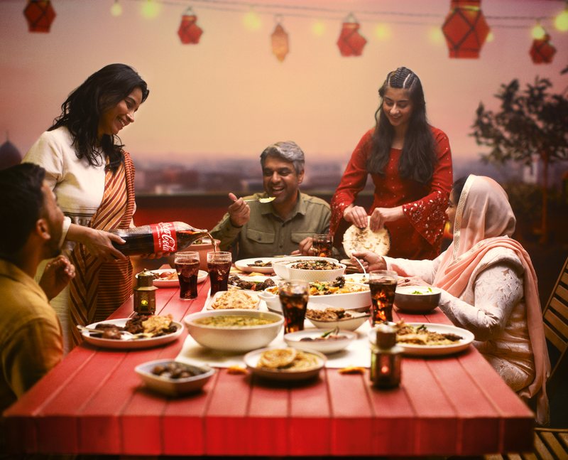 Coca-Cola Ramadan campaign image: a family sharing an evening meal at a long red table on a rooftop under hanging lanterns, with Coca-Cola being poured